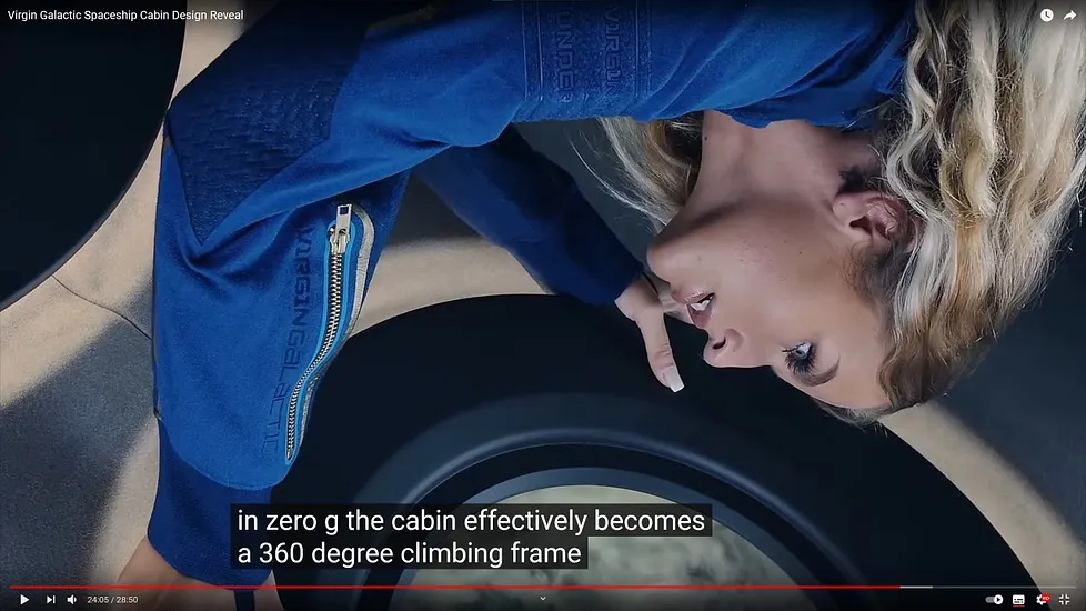 Screenshot from Virgin Galactic Spaceship Cabin Design Reveal video showing a white, blonde woman in a blue Virgin Galactic flight suit floating in zero gravity inside the spacecraft cabin. She is positioned horizontally, demonstrating weightlessness. Subtitles read: 'in zero g the cabin effectively becomes a 360 degree climbing frame'