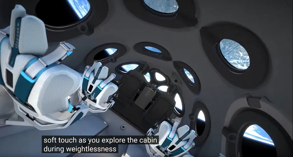 Screenshot from Virgin Galactic Spaceship Cabin Design Reveal video showing the interior of the spacecraft cabin with white and blue passenger seats visible on the left, and multiple circular windows lining the black curved wall on the right, through which Earth and sky are visible. Subtitles read: 'soft touch as you explore the cabin during weightlessness'